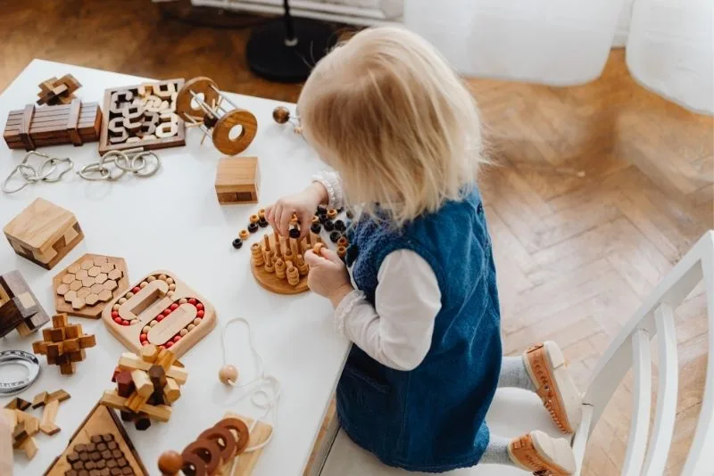 Enfant de dos, cheveux blonds, vêtu d'un gilet bleu marine et d'une chemise blanche, penché sur une table en bois garnie de jouets et puzzles en bois.