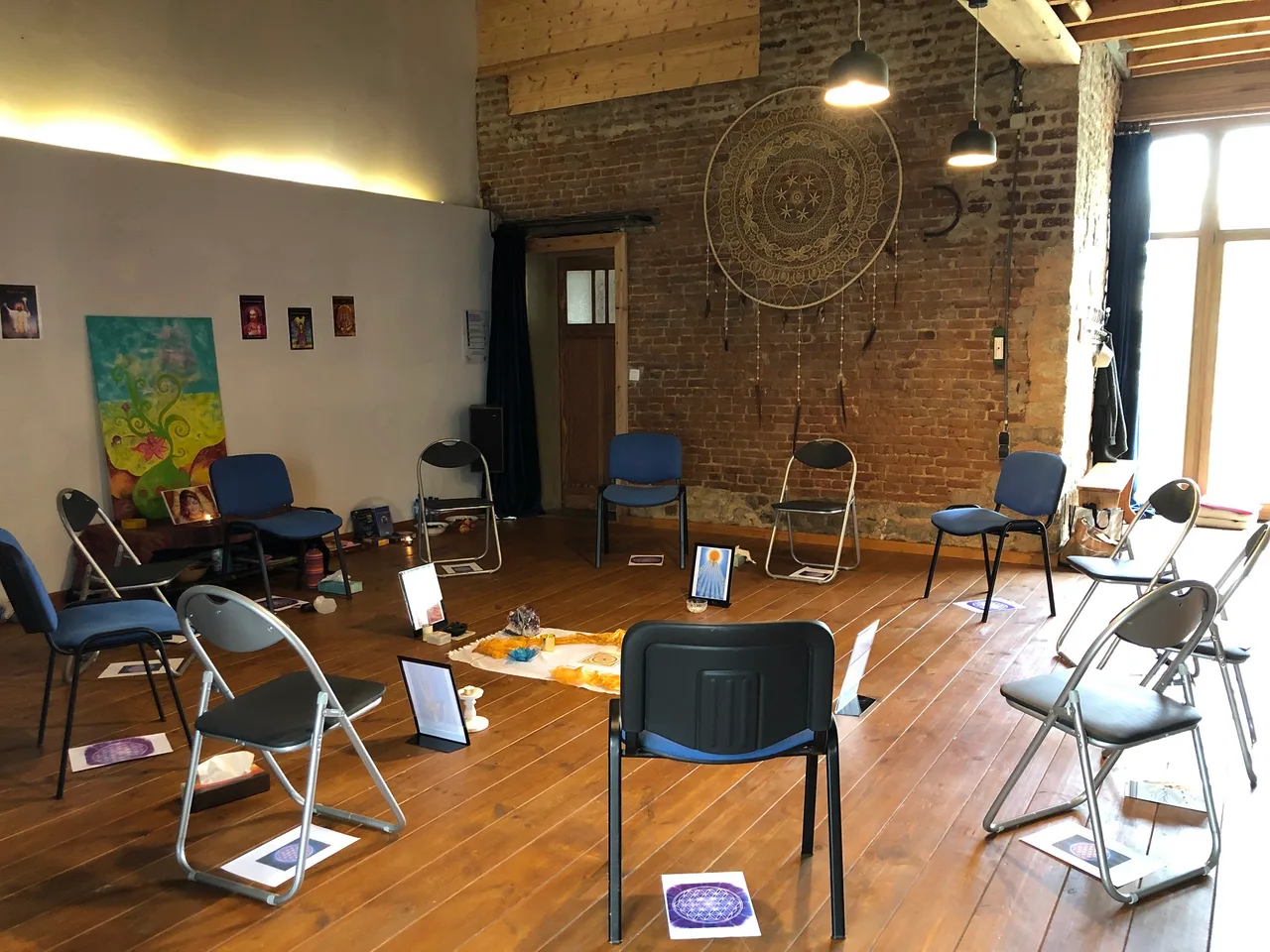 Salle de groupe avec murs en brique, chaises bleues et grises disposées en cercle, mandala doré suspendu, tableaux colorés aux murs.