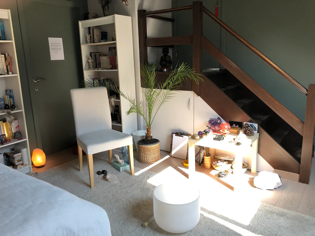 Intérieur de studio avec chaise blanche, table basse, cristaux colorés, plante, escalier en bois, étagères blanches et murs verts pâle.